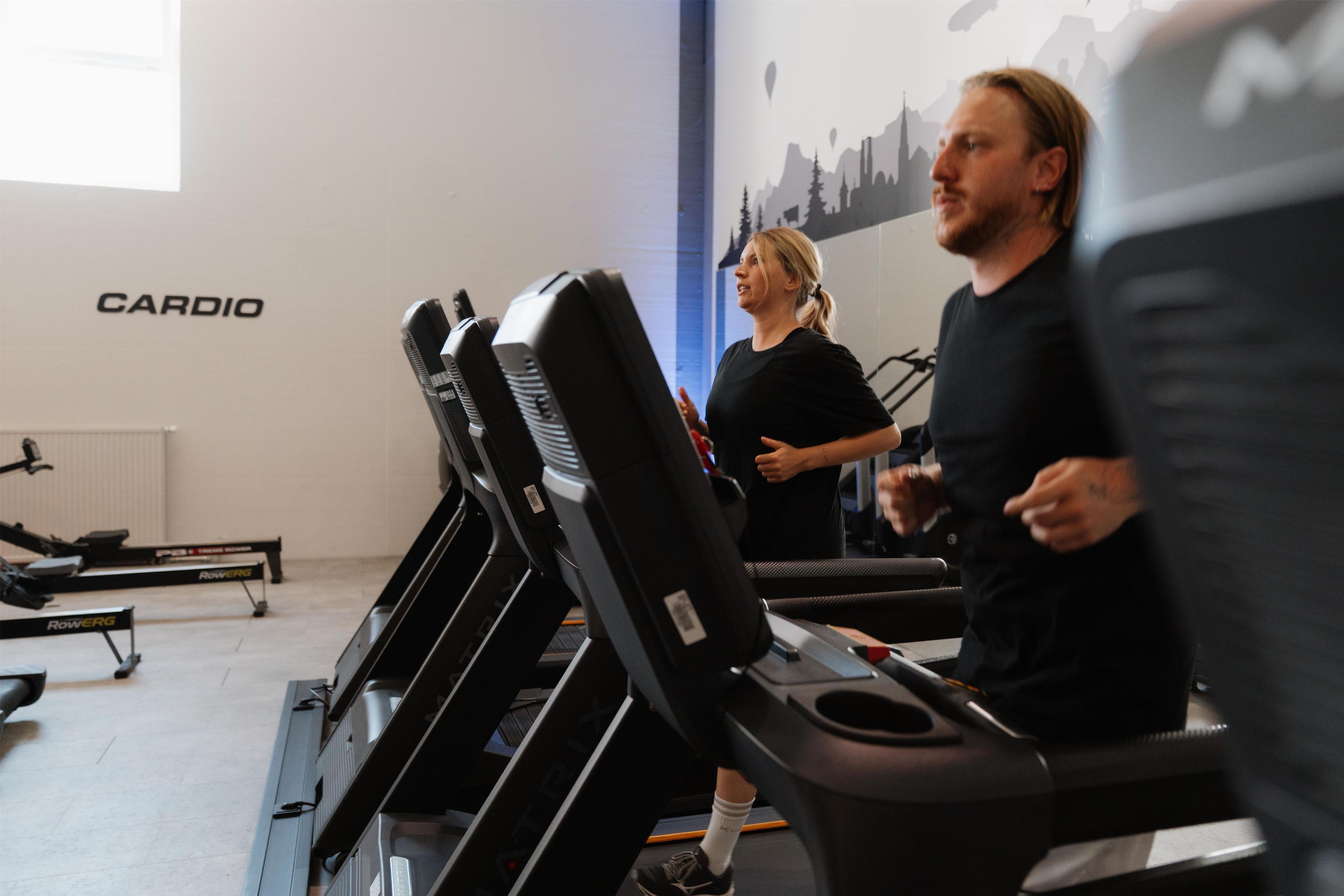 Un gimnasio con equipo de cardio, una pared donde se lee 'CARDIO' y una persona usando una caminadora.