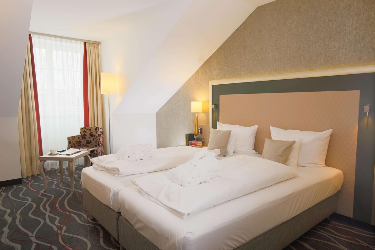 Ein Hotelzimmer mit zwei Betten, einem Sessel, einem Tisch und einer Lampe.
