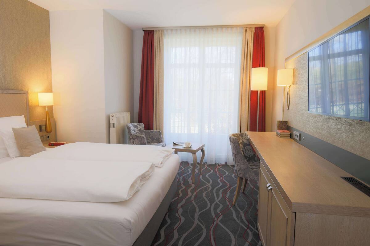 A hotel room with a large bed, a desk, two chairs, a radiator, and a large window with curtains.