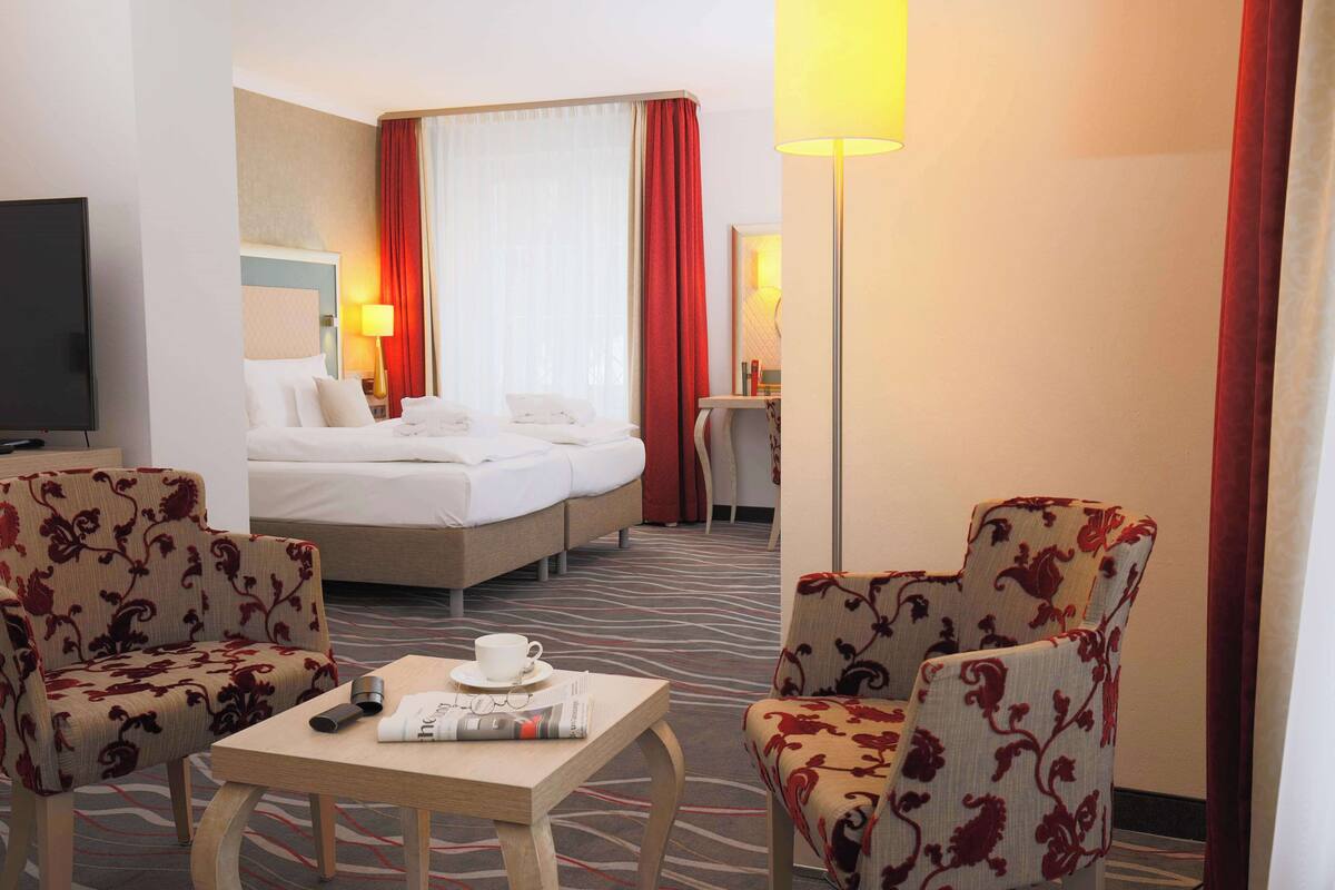 A hotel room with a bed, two armchairs, a small table with a cup and magazine, a TV, and a lamp.