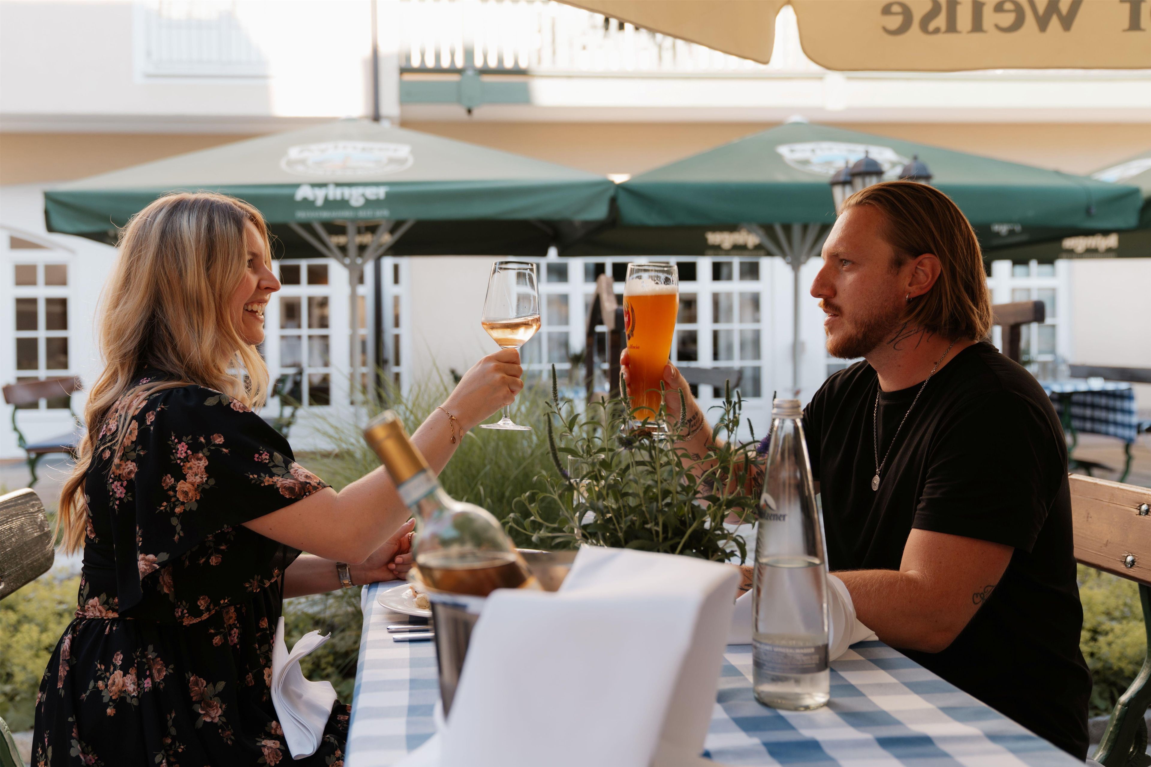 Two people toasting with drinks at an outdoor table with a checkered tablecloth, under a green umbrella.
