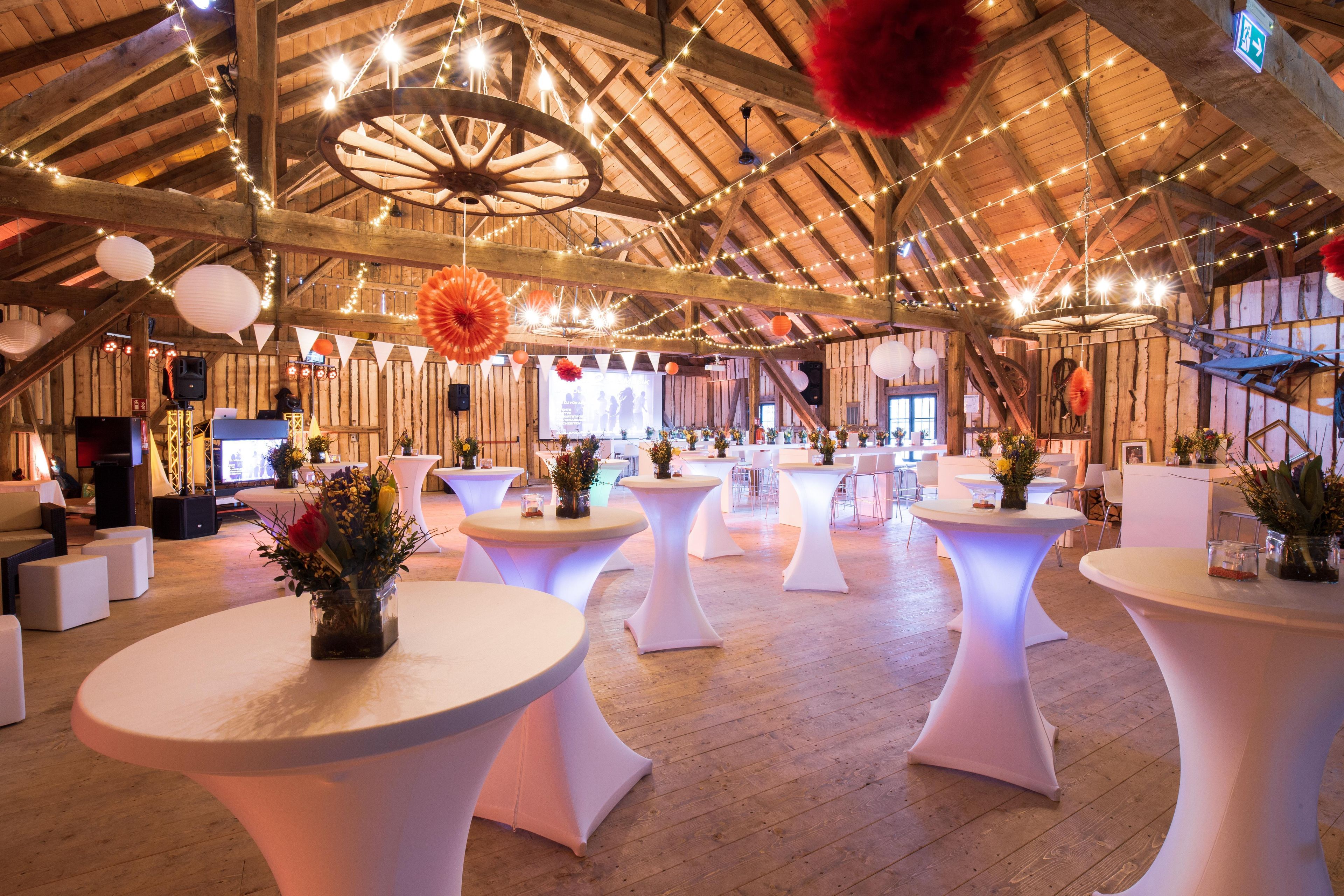 An indoor venue with multiple round tables, wooden flooring, and string lights.