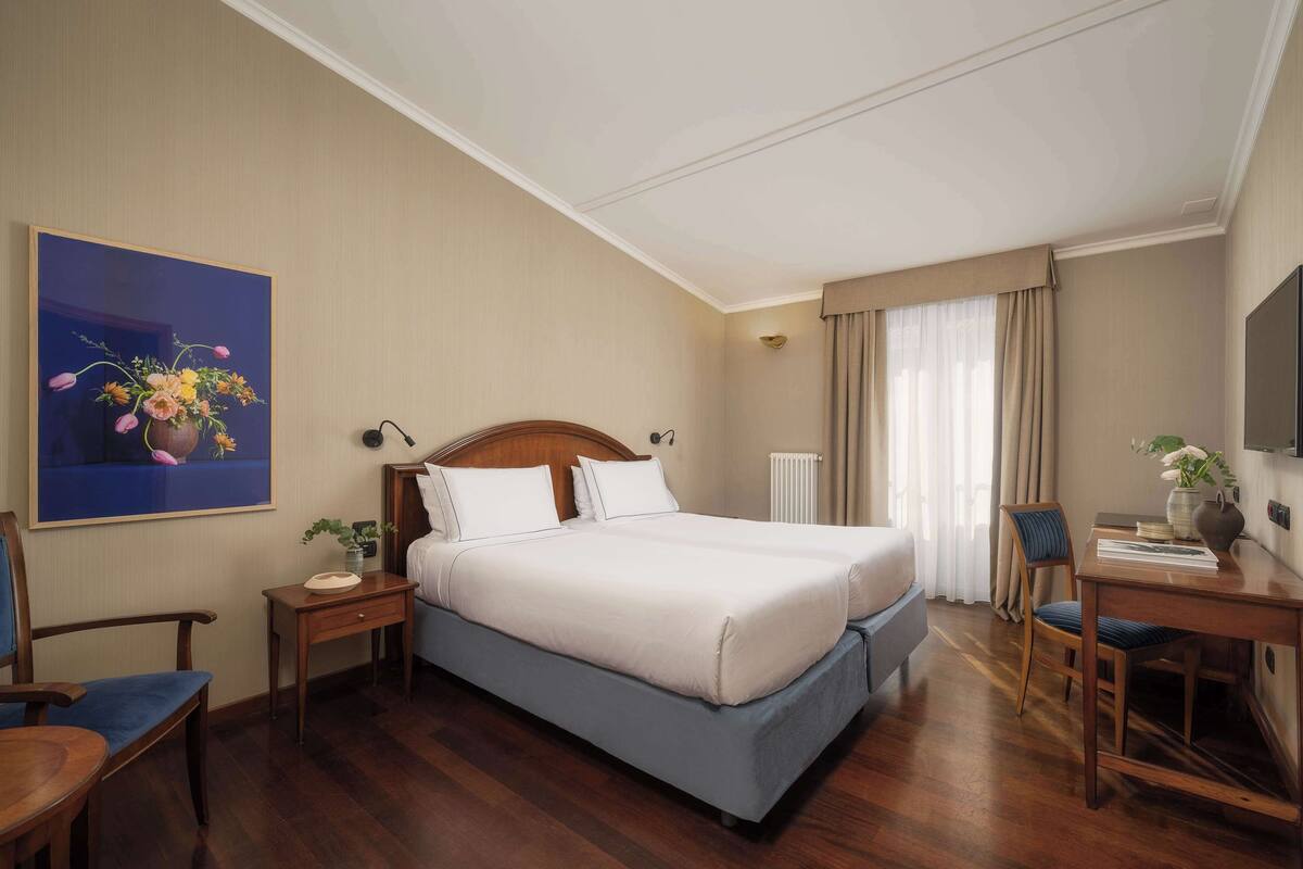 Et hotelværelse med en stor seng, et skrivebord, en stol, et lille bord, et billede og en radiator.