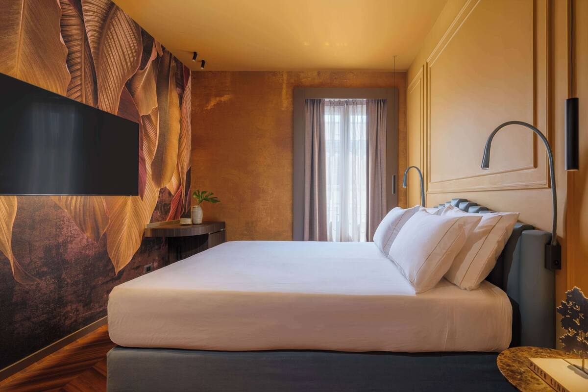 Et hotelværelse med en stor seng, et fladskærms-tv monteret på væggen, et vindue med gardiner og et moderne badeværelse med brusehoved i kurvet design.