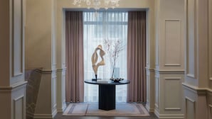 En imponerende hall med en prangende lysekrone, et rundt bord med en vase og en skulptur, samt udsigt gennem store vinduer med gardiner.