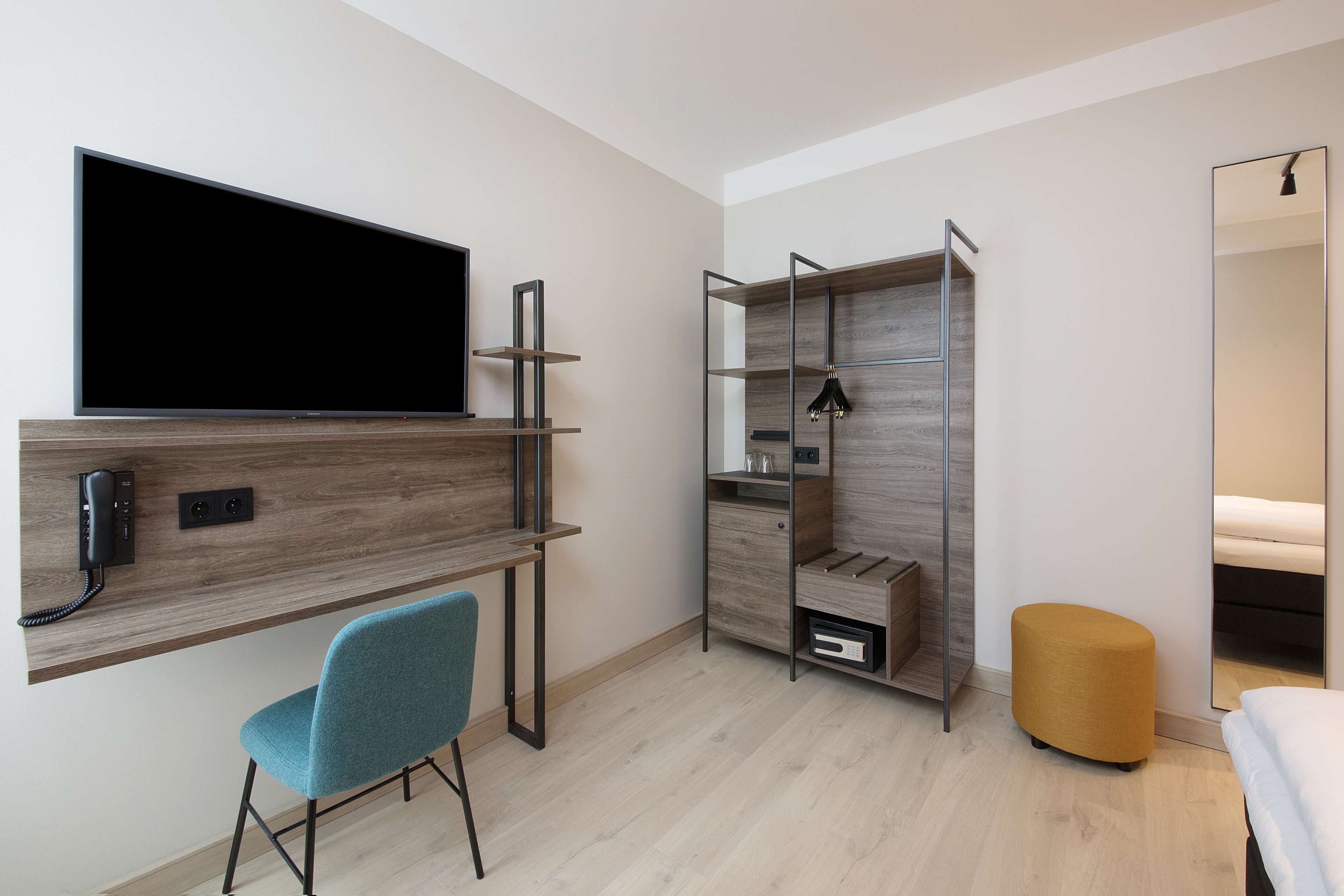Ein modernes Schlafzimmer mit an der Wand montiertem Fernseher, einem Schreibtisch mit Stuhl, einem Kleiderschrank und einem Nachttisch.