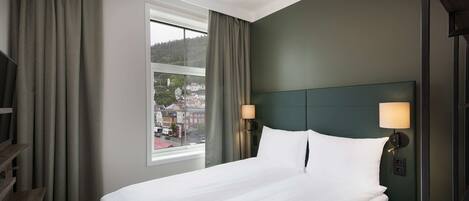 Habitación de hotel con cama, ventana con cortinas, cabecero y lámpara.