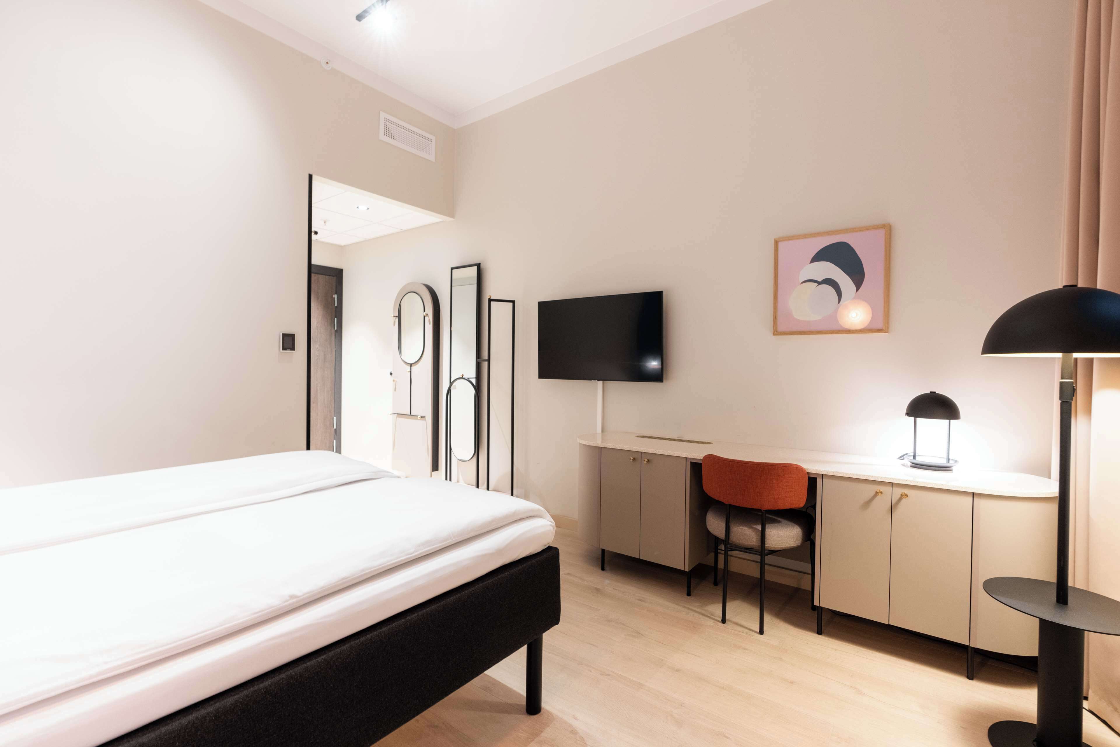 A hotel room with a bed, a desk, a chair, a TV, and a lamp.