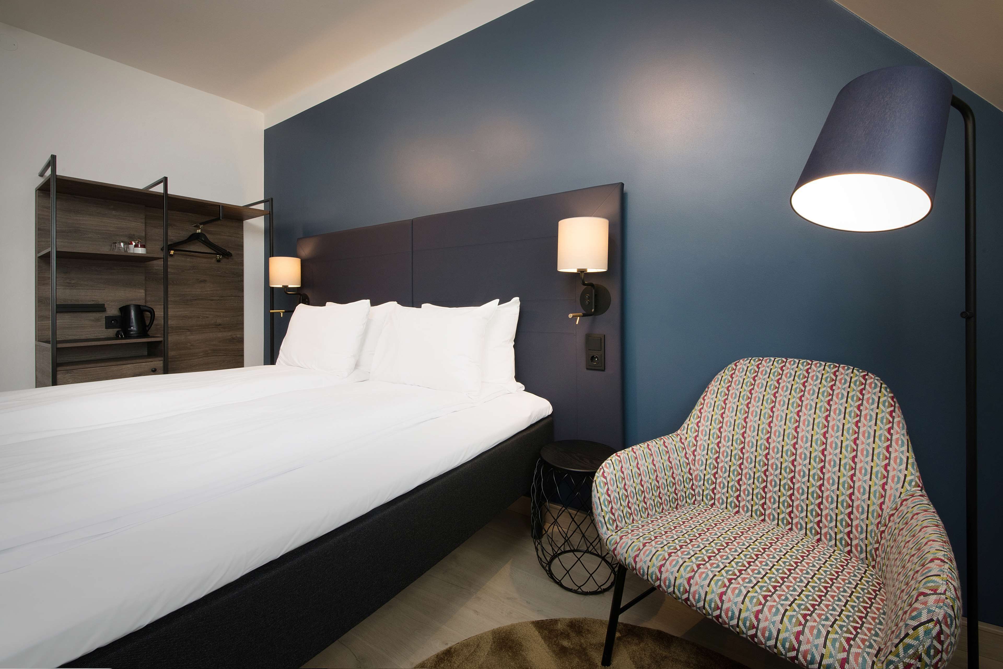 Ein ordentlich eingerichtetes Hotelzimmer mit einem großen Bett, einem Sessel, einem Nachttisch und einer Wandlampe.