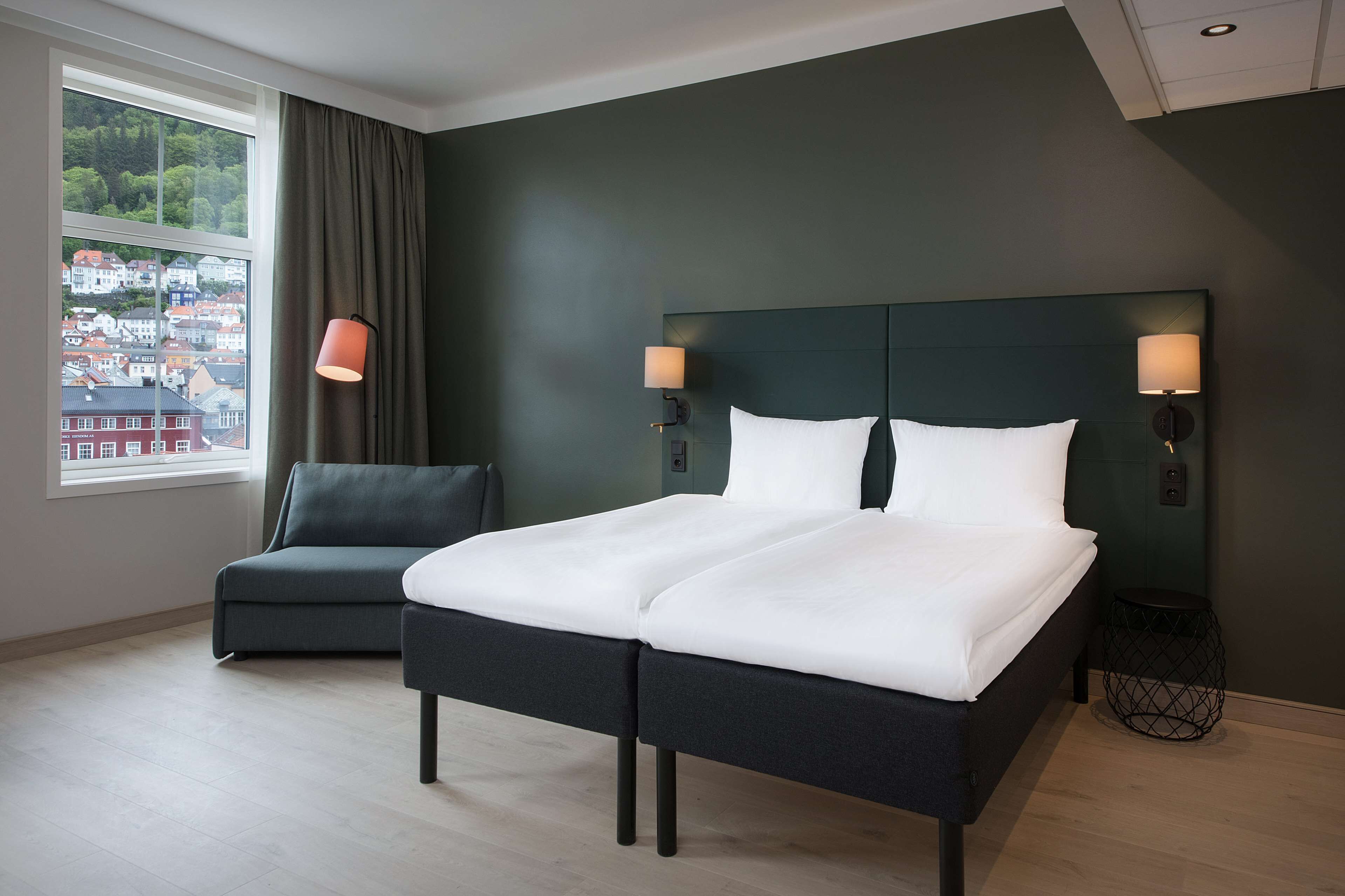 Habitación de hotel moderna con una cama grande, dos lámparas de noche, vista a un paisaje urbano a través de una ventana y un sillón oscuro.