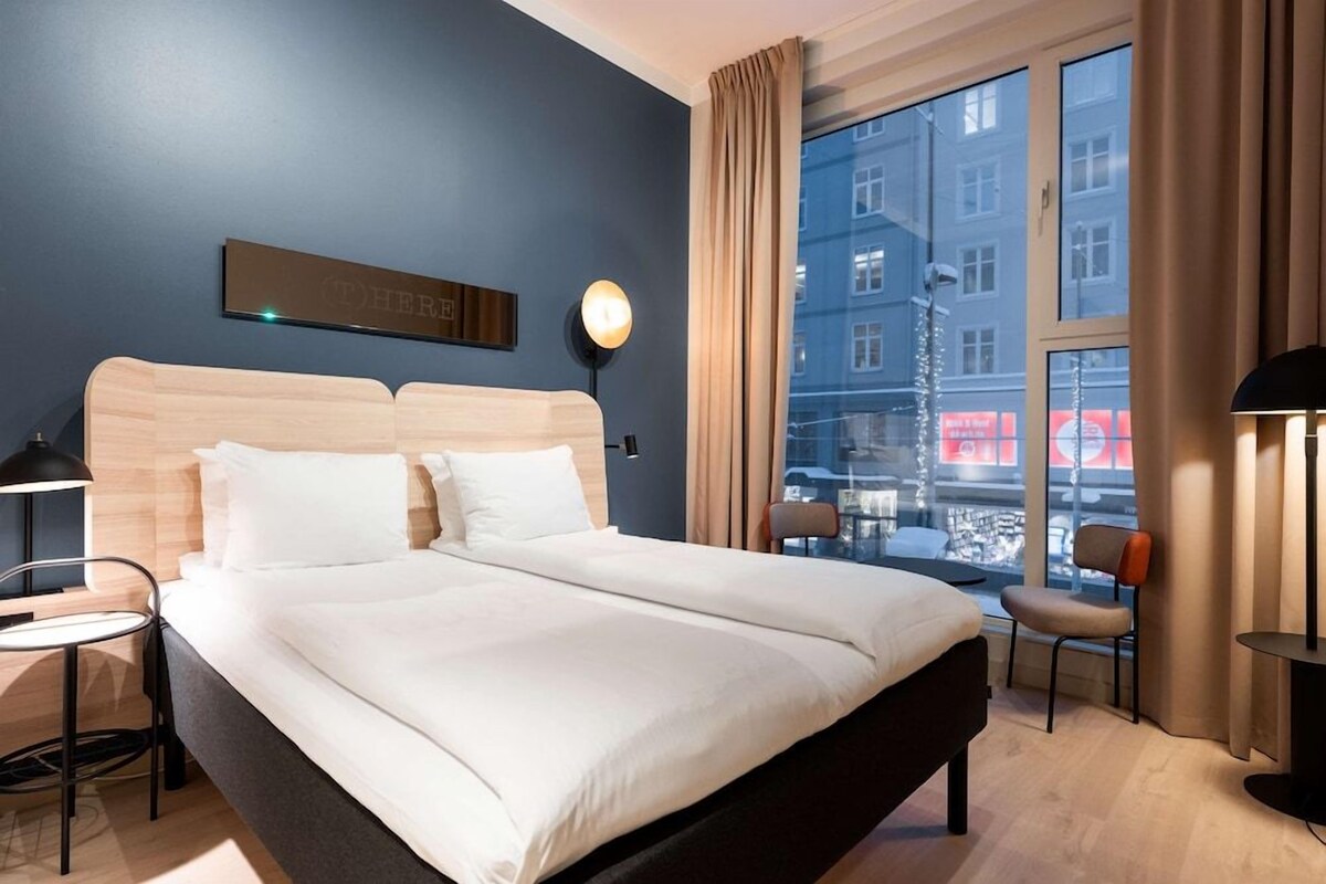 Hotellihuone, jossa on suuri sänky, yöpöytä, tuoli, ikkuna, josta on näkymä rakennuksiin, ja seinään kiinnitetty valaisin.
