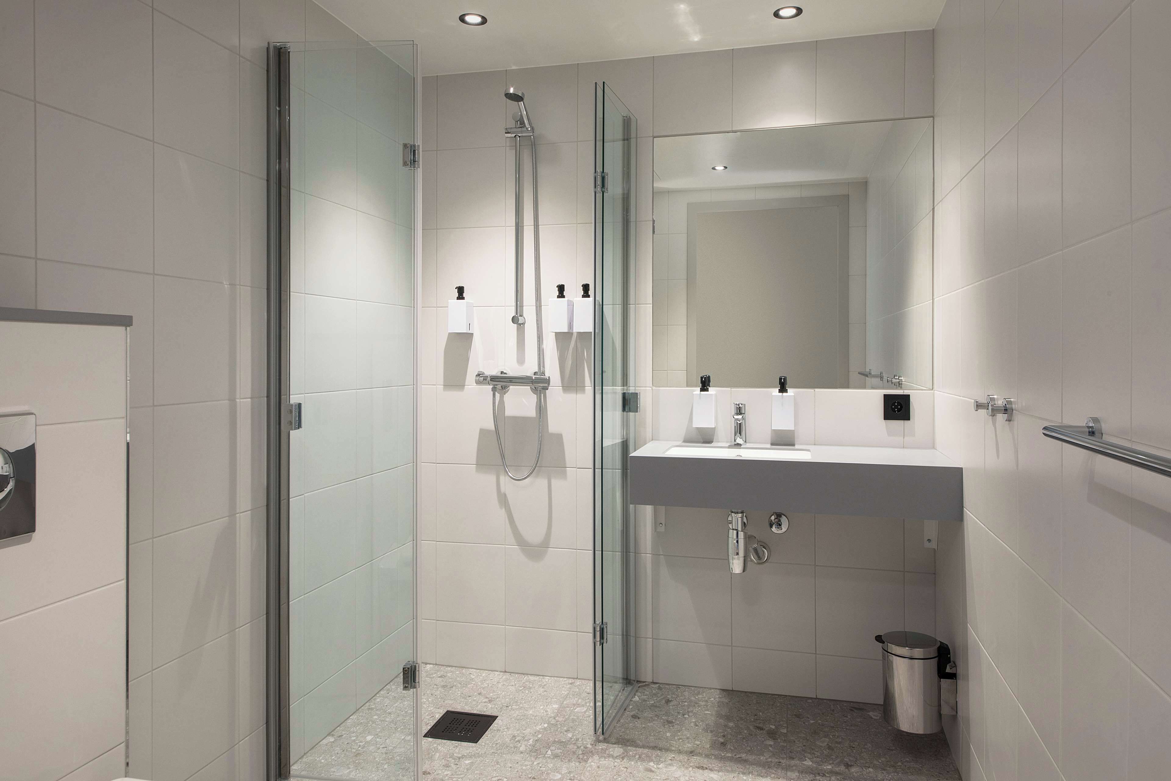 Un baño moderno con ducha de vidrio, un lavabo blanco con un espejo encima y un pequeño cubo de basura.