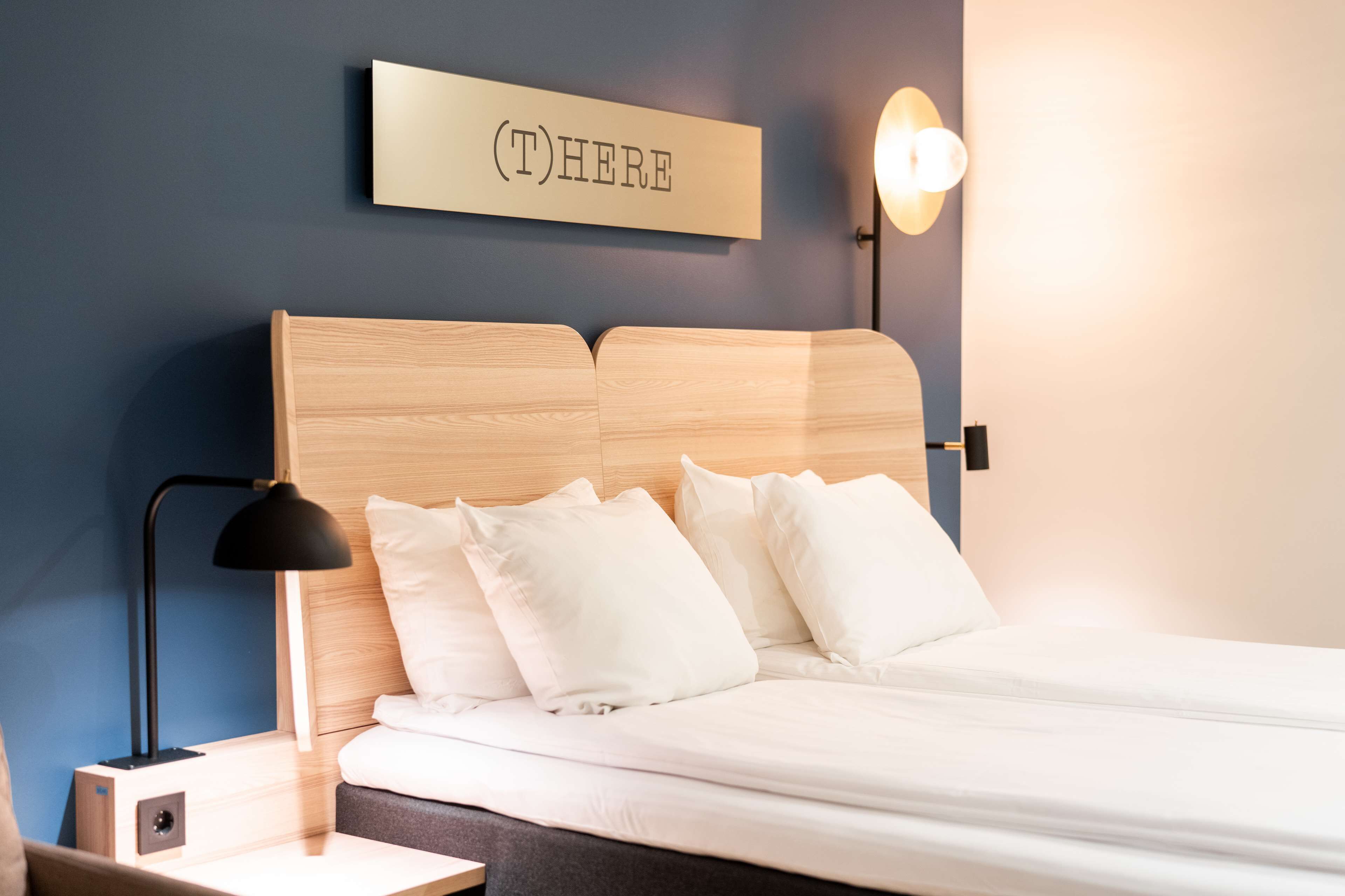A neatly made bed with white linens, a wooden headboard, and a bedside lamp. A sign above the bed reads "(T) HERE".