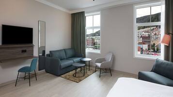 A modern hotel room with a large bed, two sofas, a TV, and a city view.