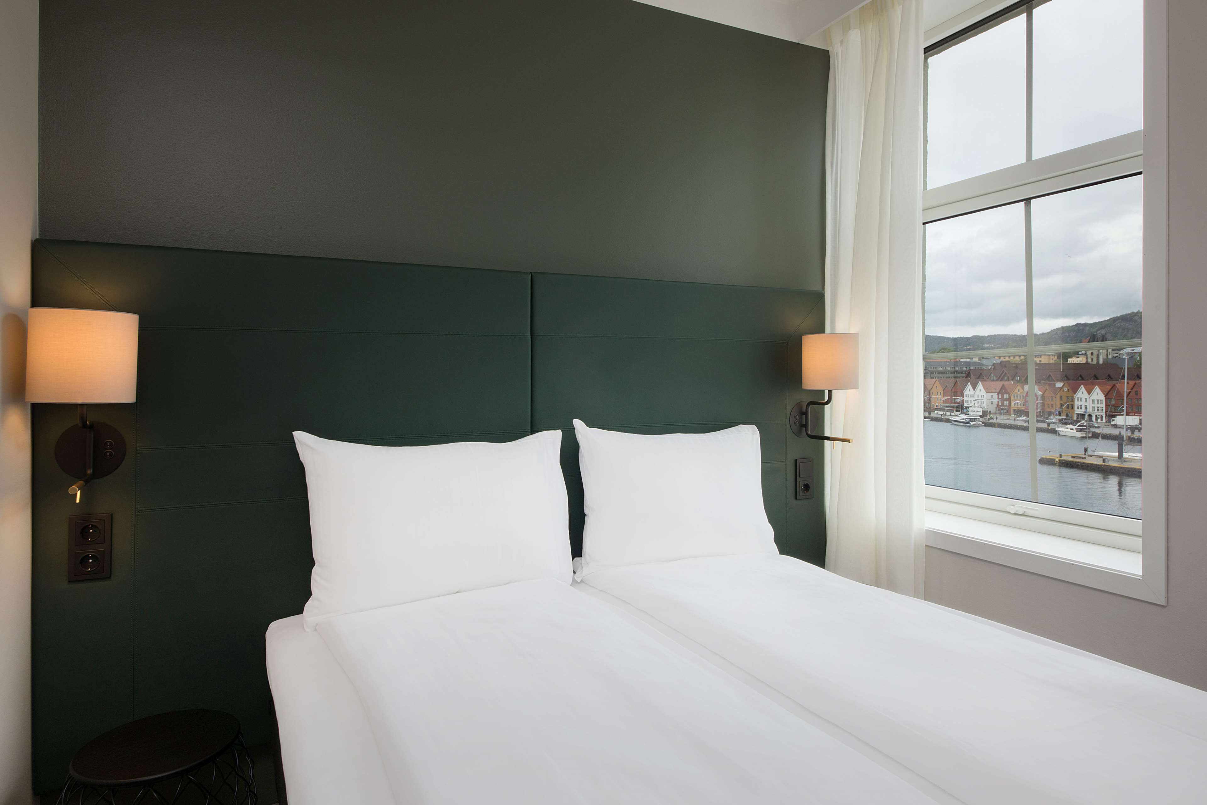 Un lit bien fait, avec du linge de lit blanc, une tête de lit vert foncé et deux lampes fixées au mur.