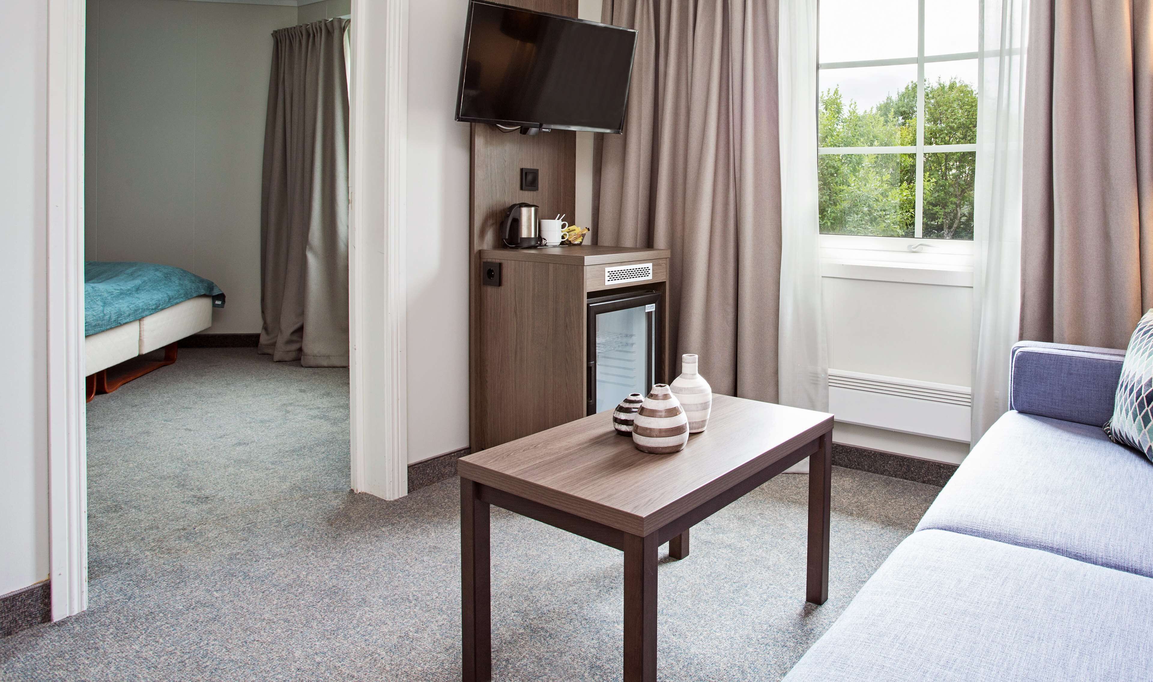 Habitación de hotel con cama, televisor montado en la pared, una pequeña mesa con adornos, un sofá y una ventana con cortinas.