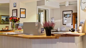 A reception desk with a laptop, a potted plant, and various informational posters. - Scandic Leknes Lofoten (Vestvågøy)