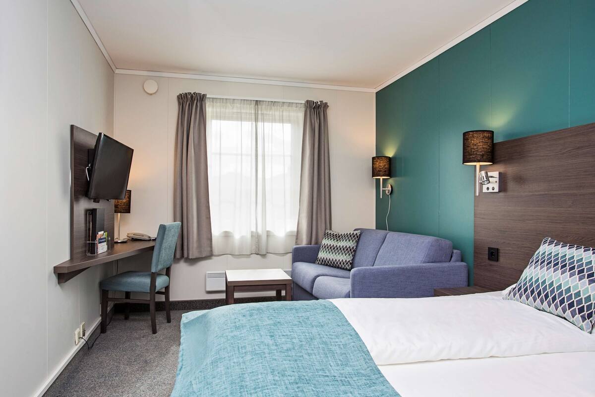 A hotel room with a bed, a desk with a chair, a television, a sofa, and two wall-mounted lamps.