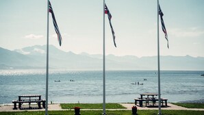 Trois mâts sur lesquels flottent des drapeaux, un banc et une table de pique-nique, le tout situé au bord d’une étendue d’eau, avec des montagnes en arrière-plan.