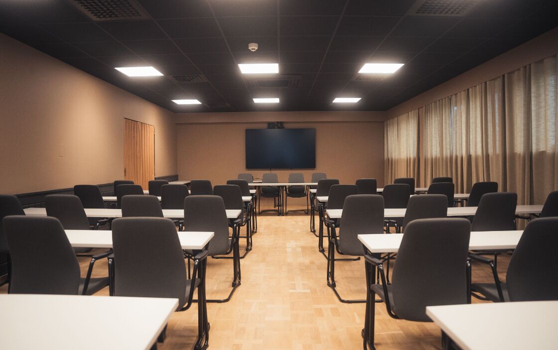Ett konferensrum med rader av bord och stolar, en stor skärm längst fram och ett fönster med gardiner.