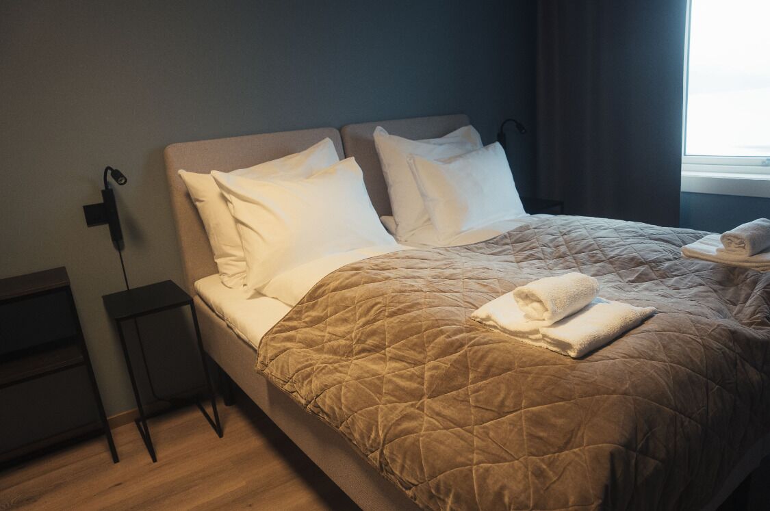 A neatly made bed with a quilted brown cover, white pillows, and a folded towel on the side.