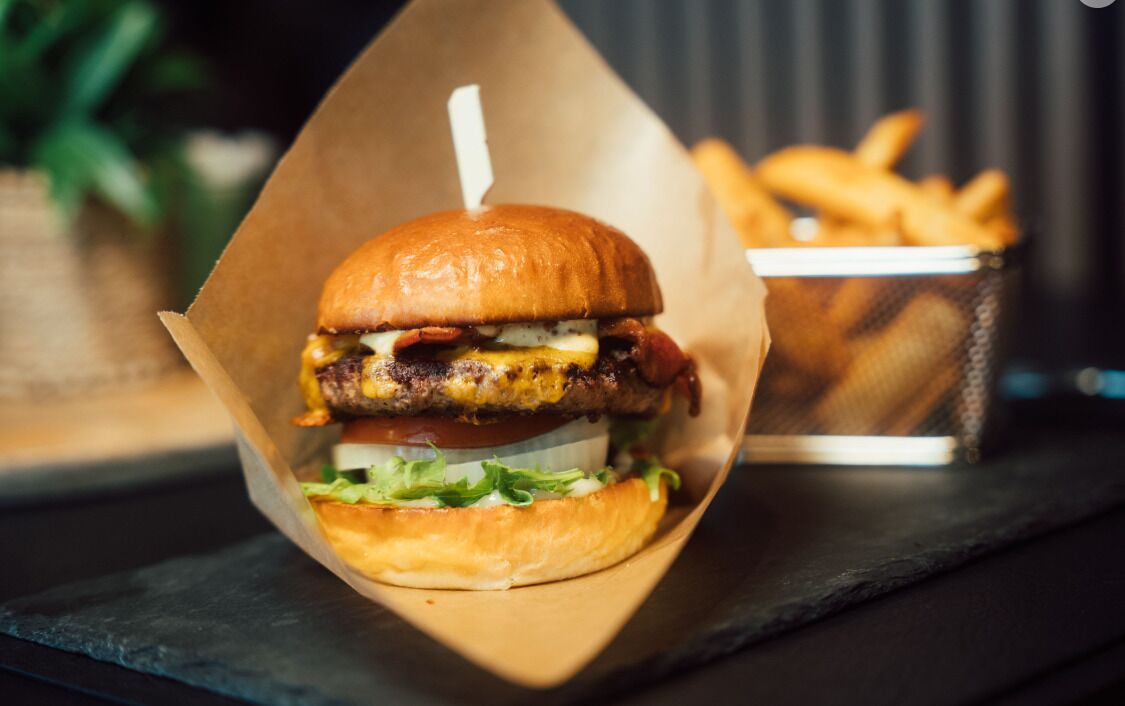 A burger in a paper container with fries in the background.