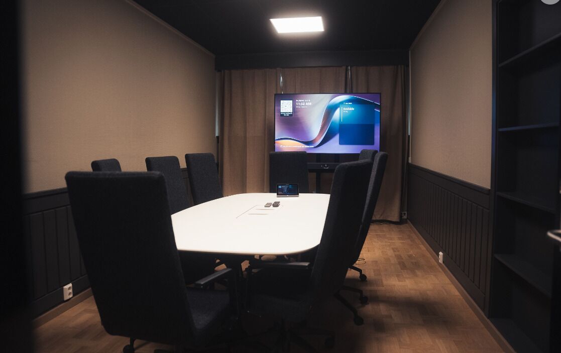 A conference room with a white table, six black chairs, a large screen, and a wooden floor.