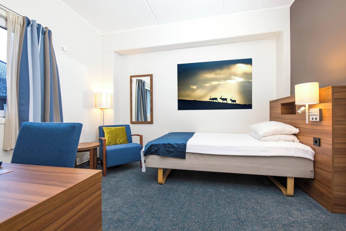 A hotel room with a bed, a desk, a chair, a lamp, and a painting on the wall.