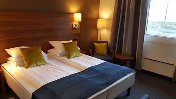 Habitación de hotel con cabecera de madera, cama con sábanas blancas y dos almohadas verdes, una mesita con una silla y una ventana con cortinas.
