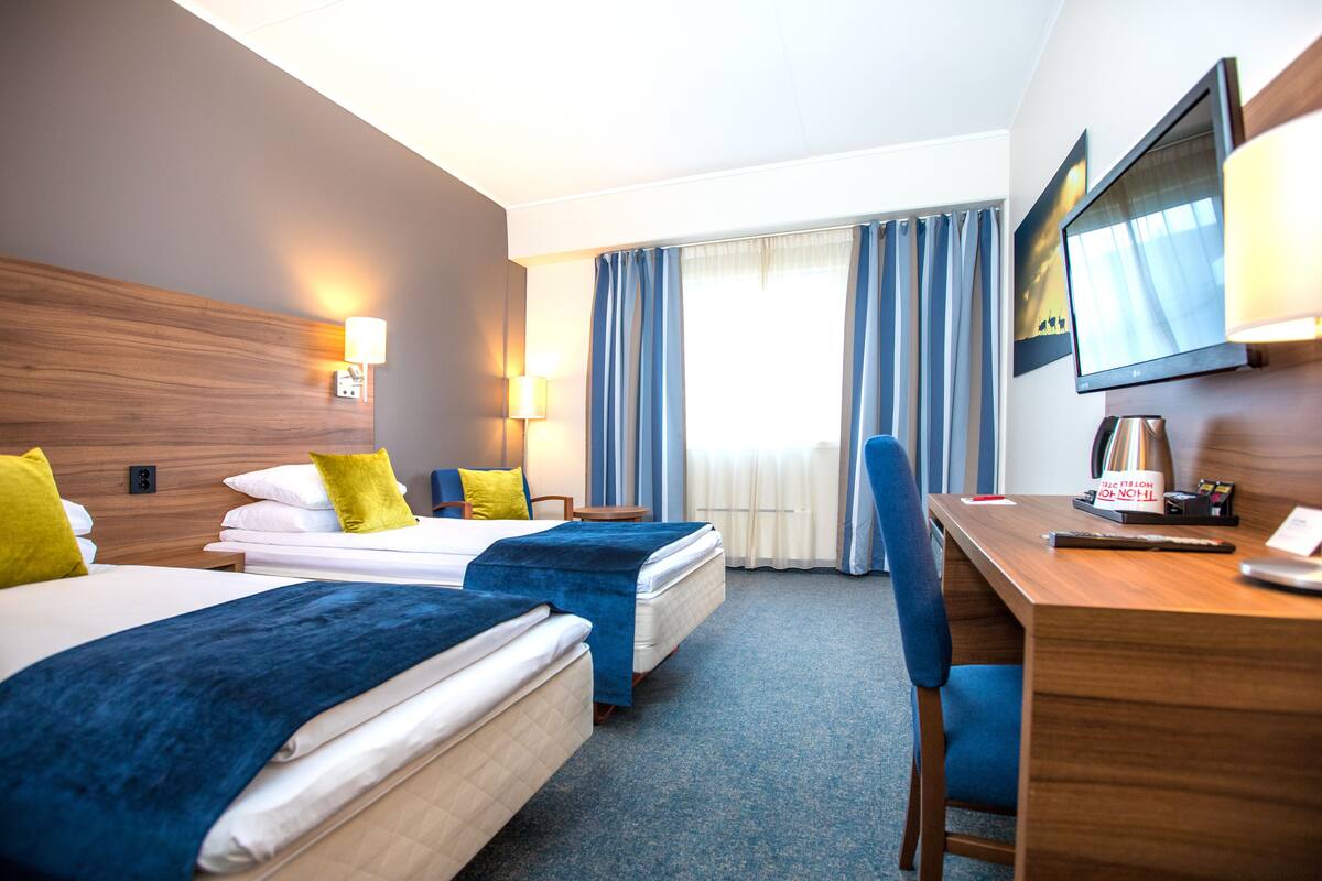 A hotel room with two beds, a desk, a chair, a TV, and a window with curtains.