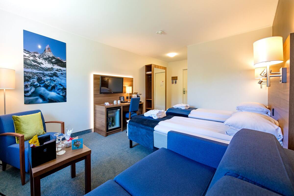 A hotel room with two beds, a desk, a chair, a TV, and a painting of a mountain scene.