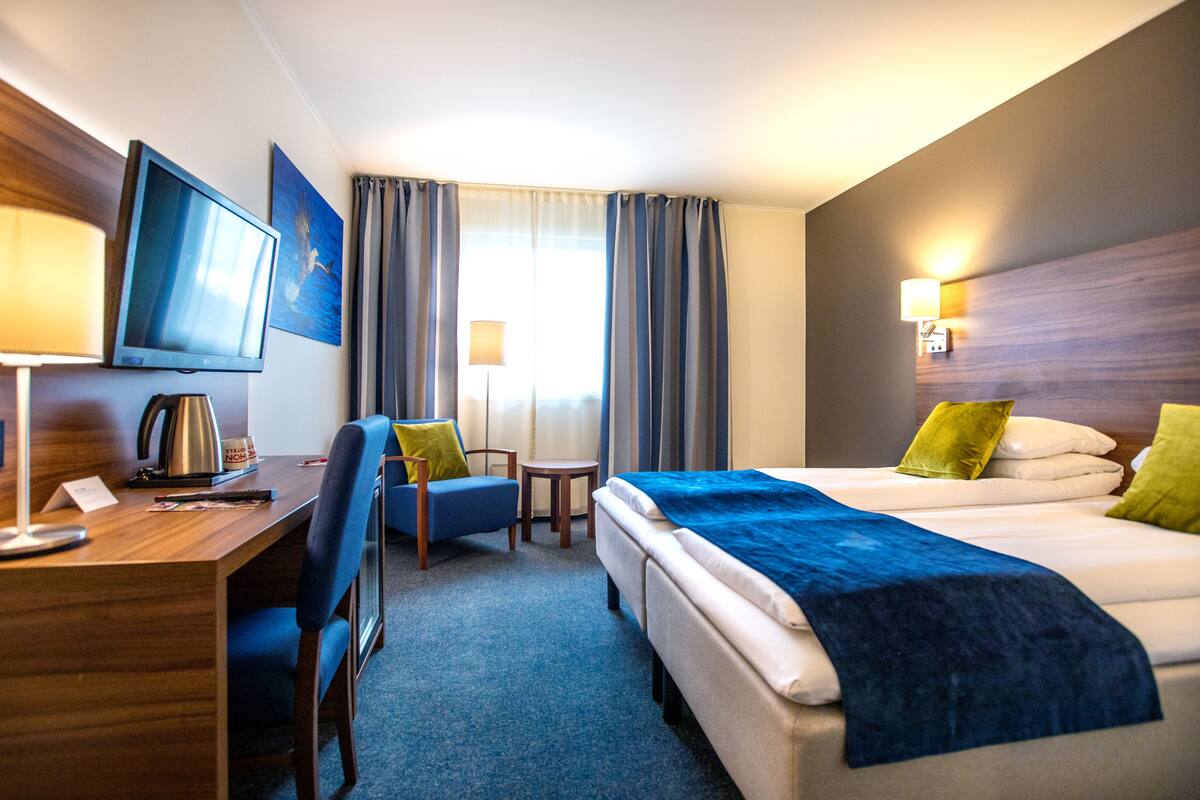 A hotel room with a bed, a desk with a computer, a chair, a television, and a window with curtains.