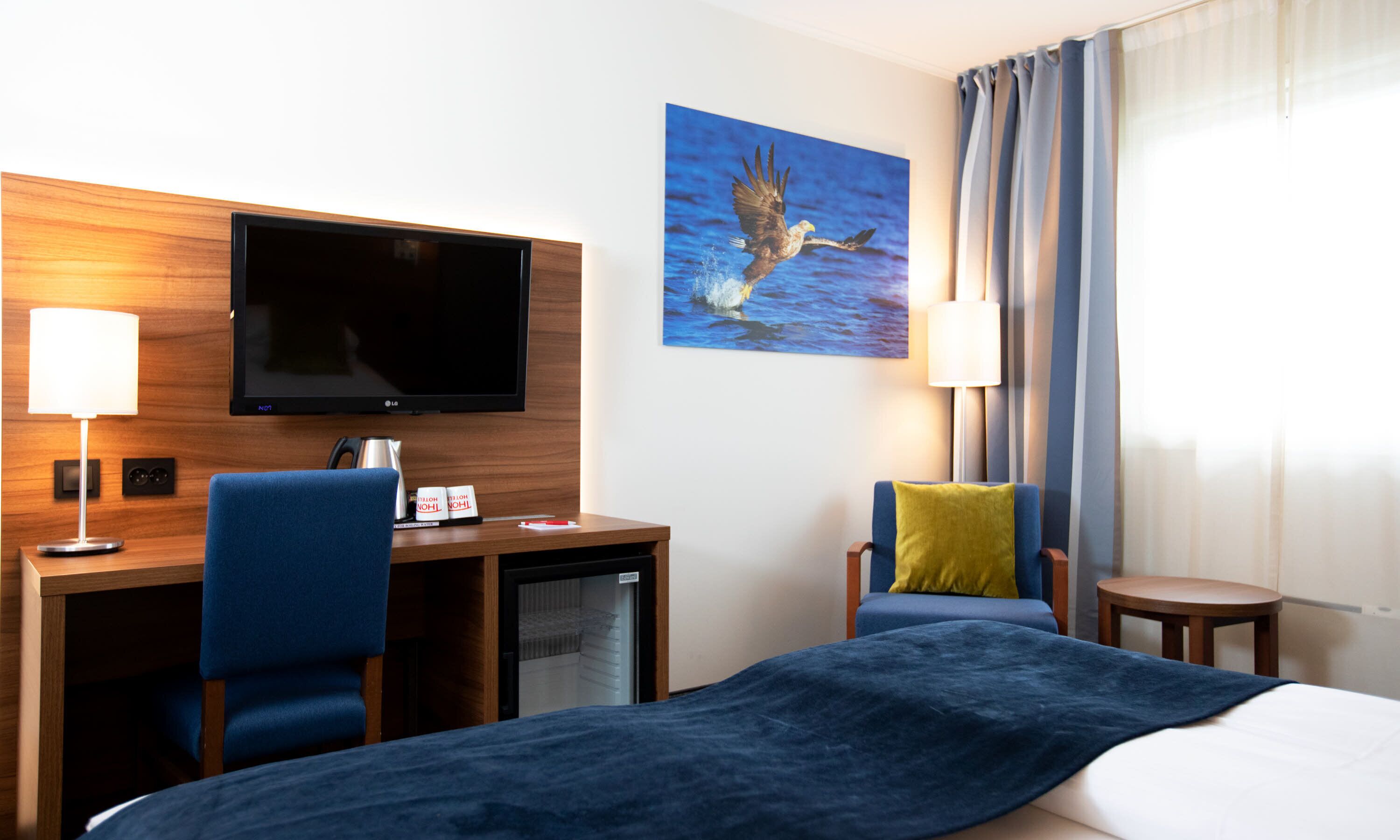 A hotel room with a bed, a desk with a TV, two chairs, a lamp, and a painting of a bird.