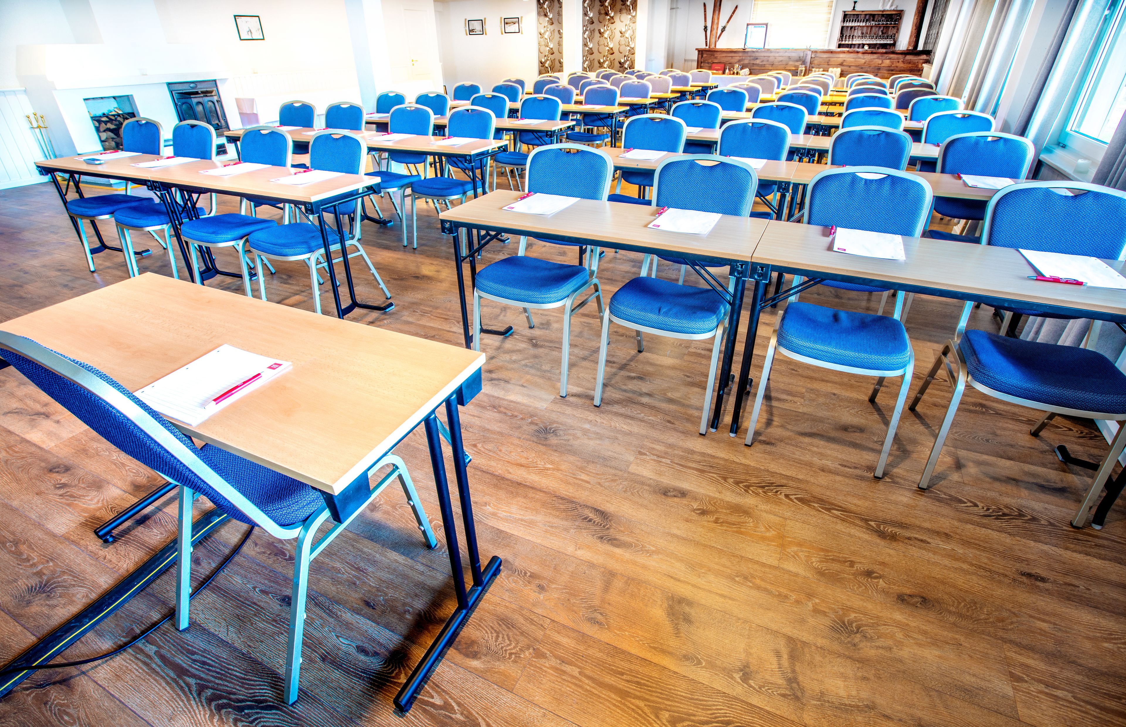 A classroom with rows of desks and chairs, wooden floors, and a fireplace in the background.
