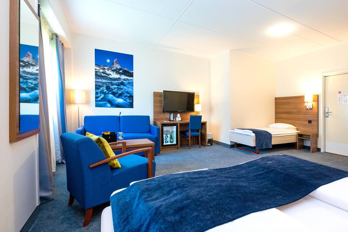 A hotel room with a bed, a desk with a computer, a chair, a sofa, a TV, and a large window.
