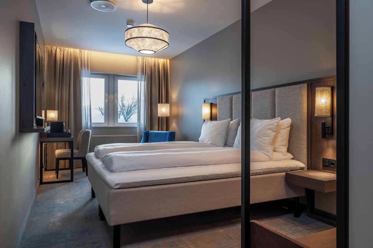 Quarto de hotel com cama, mesas de cabeceira, escrivaninha, cadeira, janela com cortinas e luminária pendente.