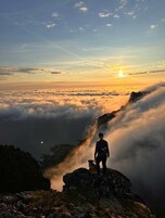 Una persona parada sobre un saliente rocoso contemplando un extenso paisaje cubierto de nubes con un atardecer de fondo.