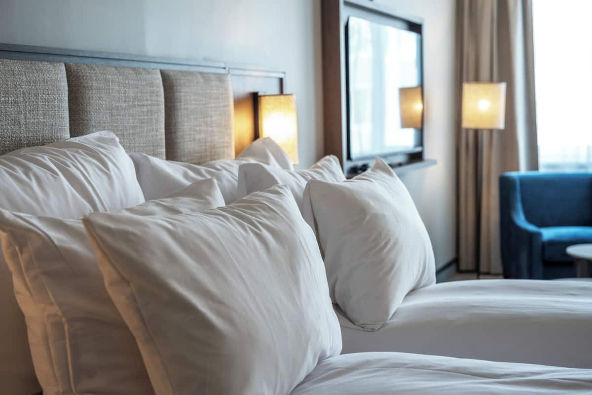 Quarto de hotel com cama, almofadas, cabeceira, luminária, cadeira e janela.