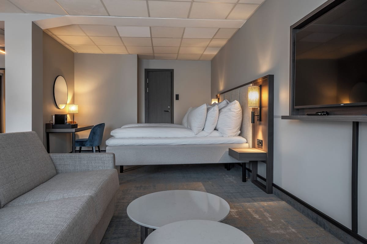 Quarto de hotel moderno com sofá, cama com roupa de cama branca, televisão de tela plana e escrivaninha com abajur.
