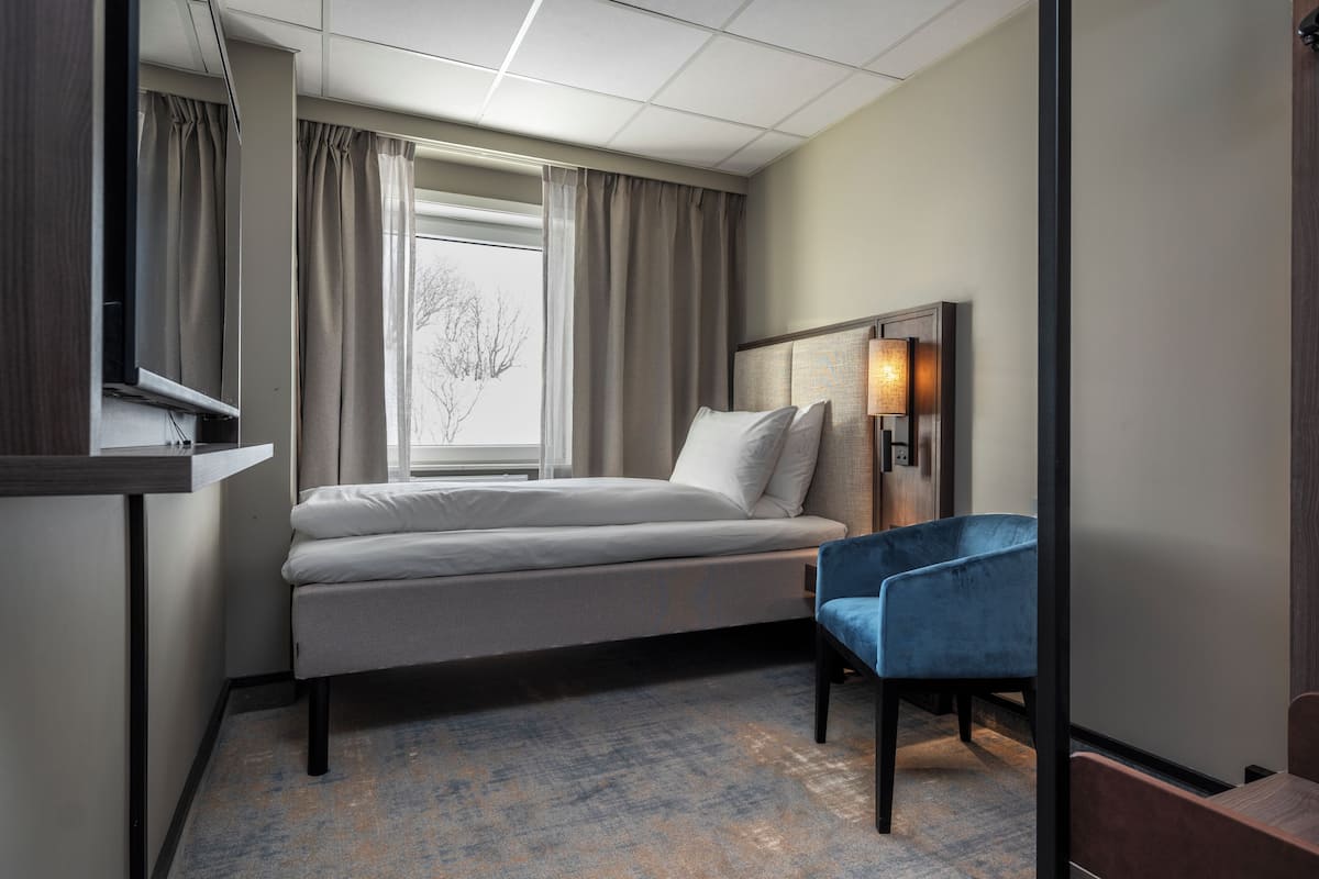 Quarto de hotel com cama, escrivaninha, cadeira, luminária e janela com cortinas.