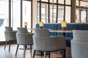 Un área de descanso con sillas tapizadas en gris y un sofá azul, una mesa redonda con una lámpara, y ventanas grandes que ofrecen vistas al exterior.