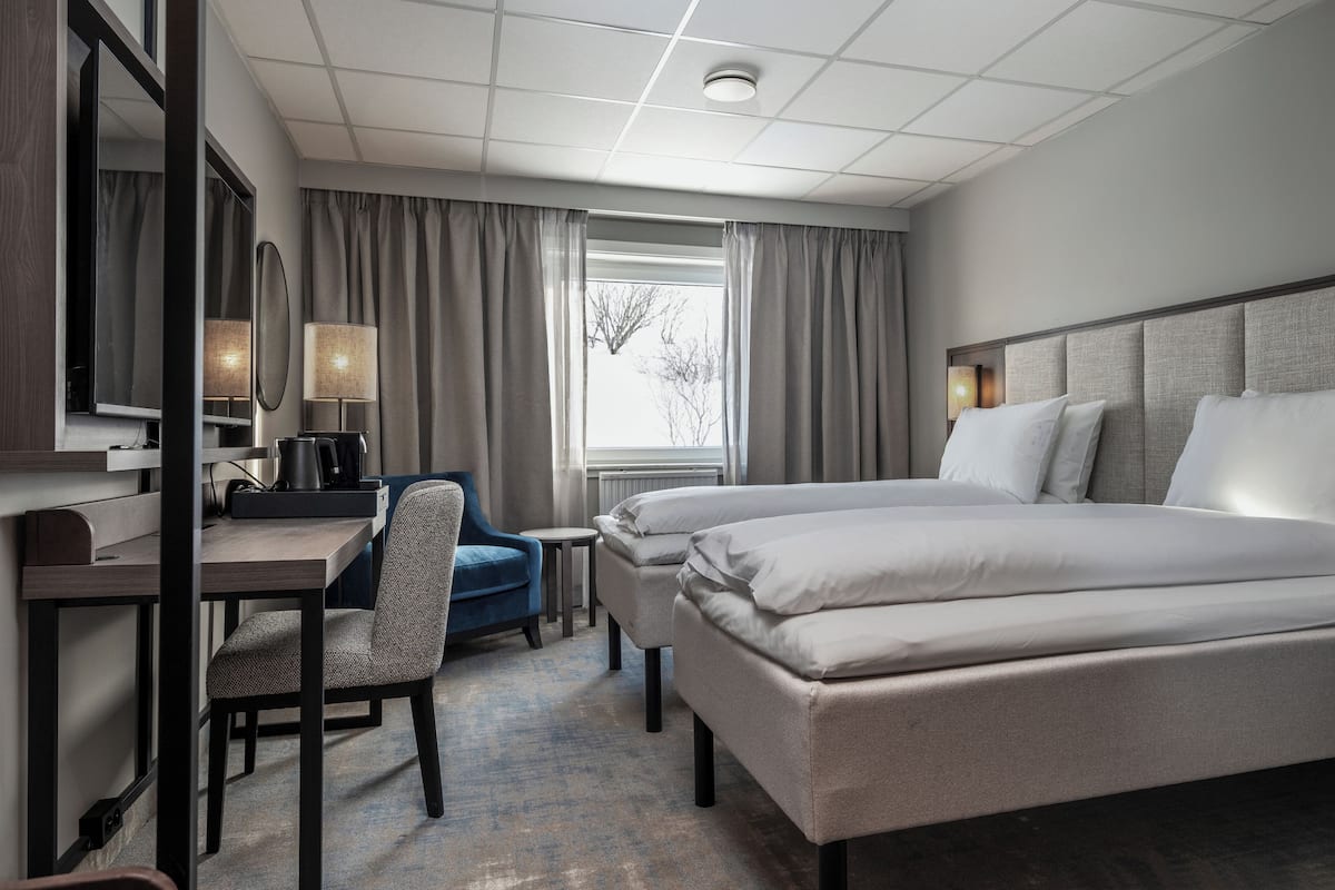 Quarto de hotel com cama, escrivaninha e cadeira, mesinha, abajur e janela com cortinas.