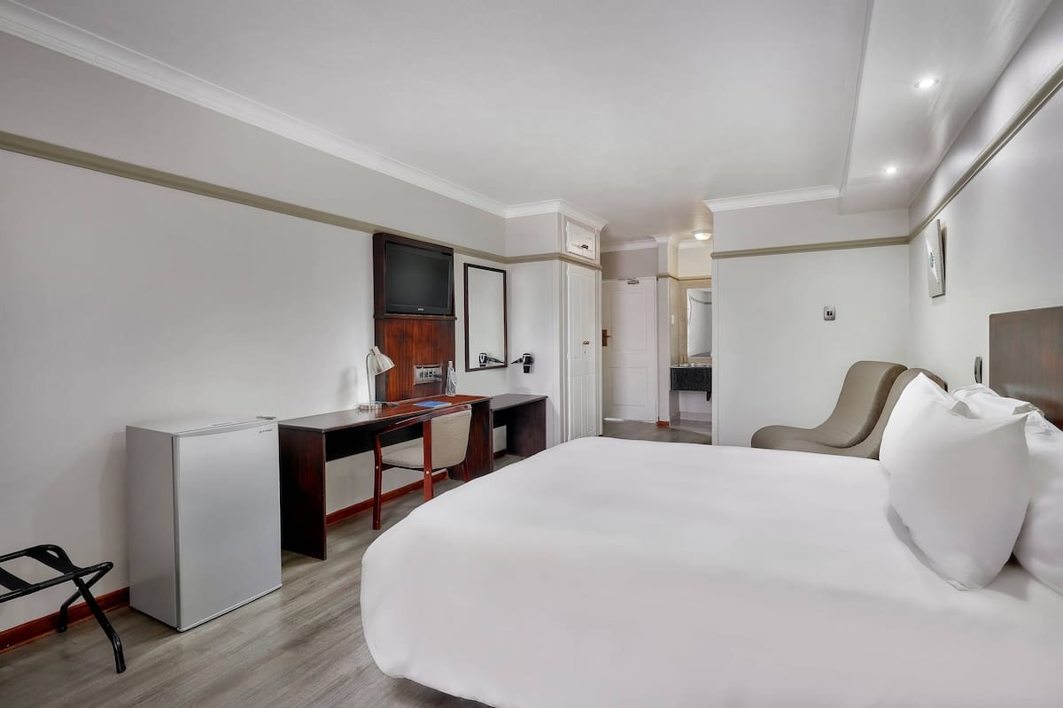 Une chambre d’hôtel avec un grand lit, un bureau avec une chaise, un petit réfrigérateur, une télévision et une porte donnant sur une salle de bain.