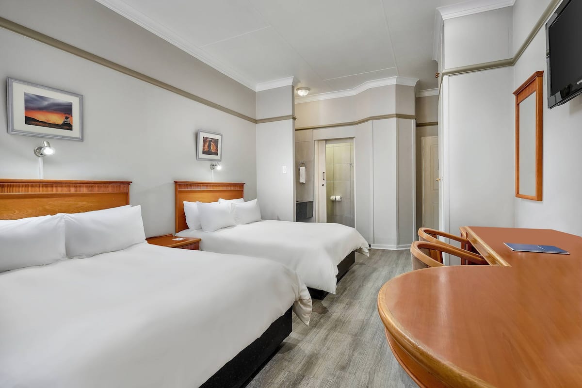 Une chambre d’hôtel avec deux lits, un bureau en bois, une télévision et une porte donnant sur une salle de bain.