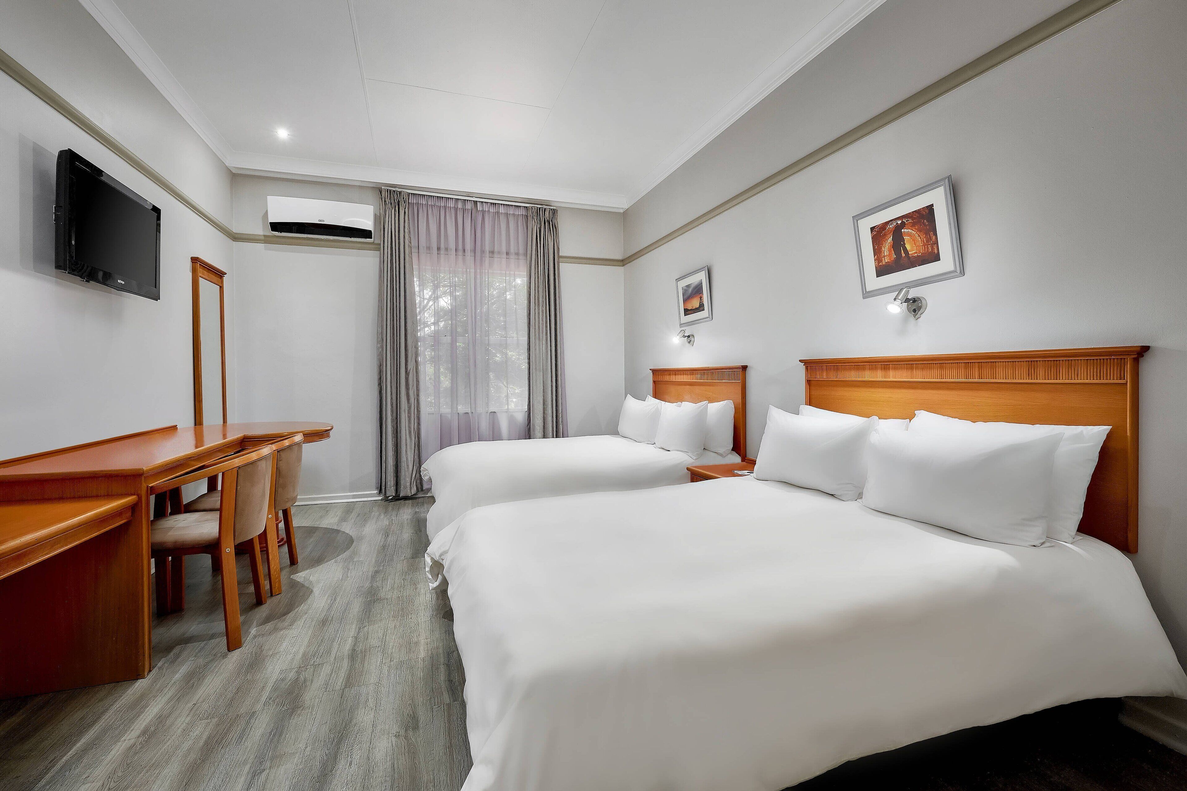 Ein Hotelzimmer mit zwei Betten, einem Schreibtisch, einem Stuhl, einem Fernseher und einem Fenster mit Vorhängen.