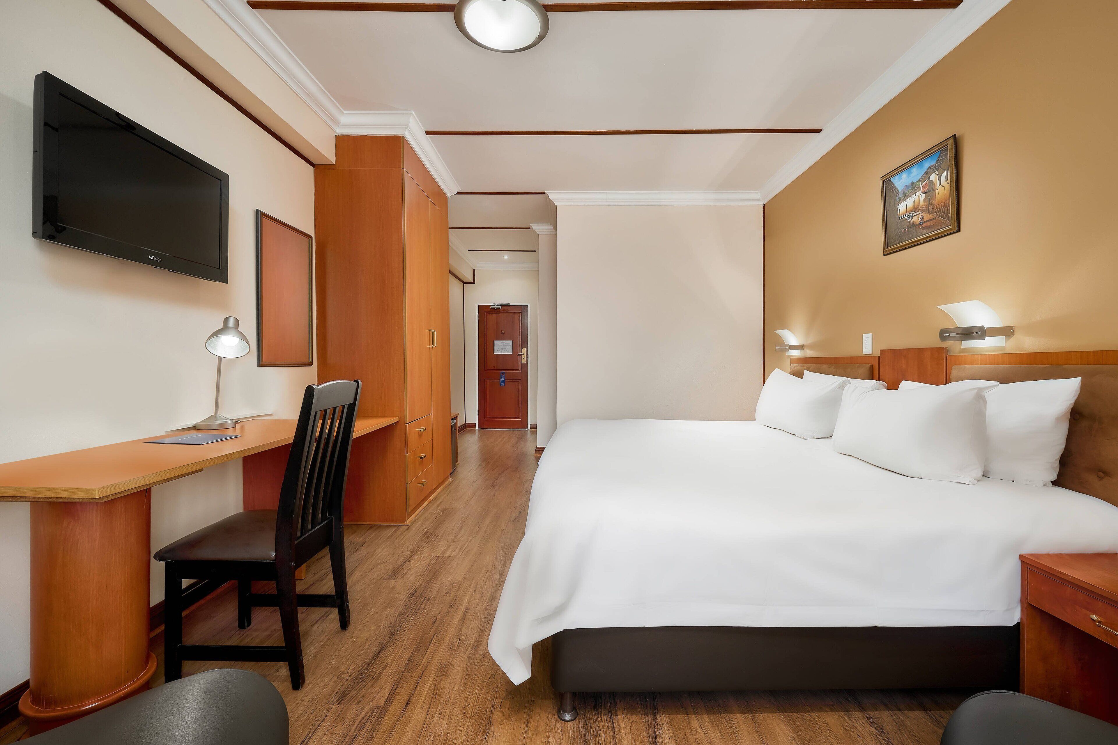 A hotel room with a bed, desk, chair, TV, and a painting on the wall.