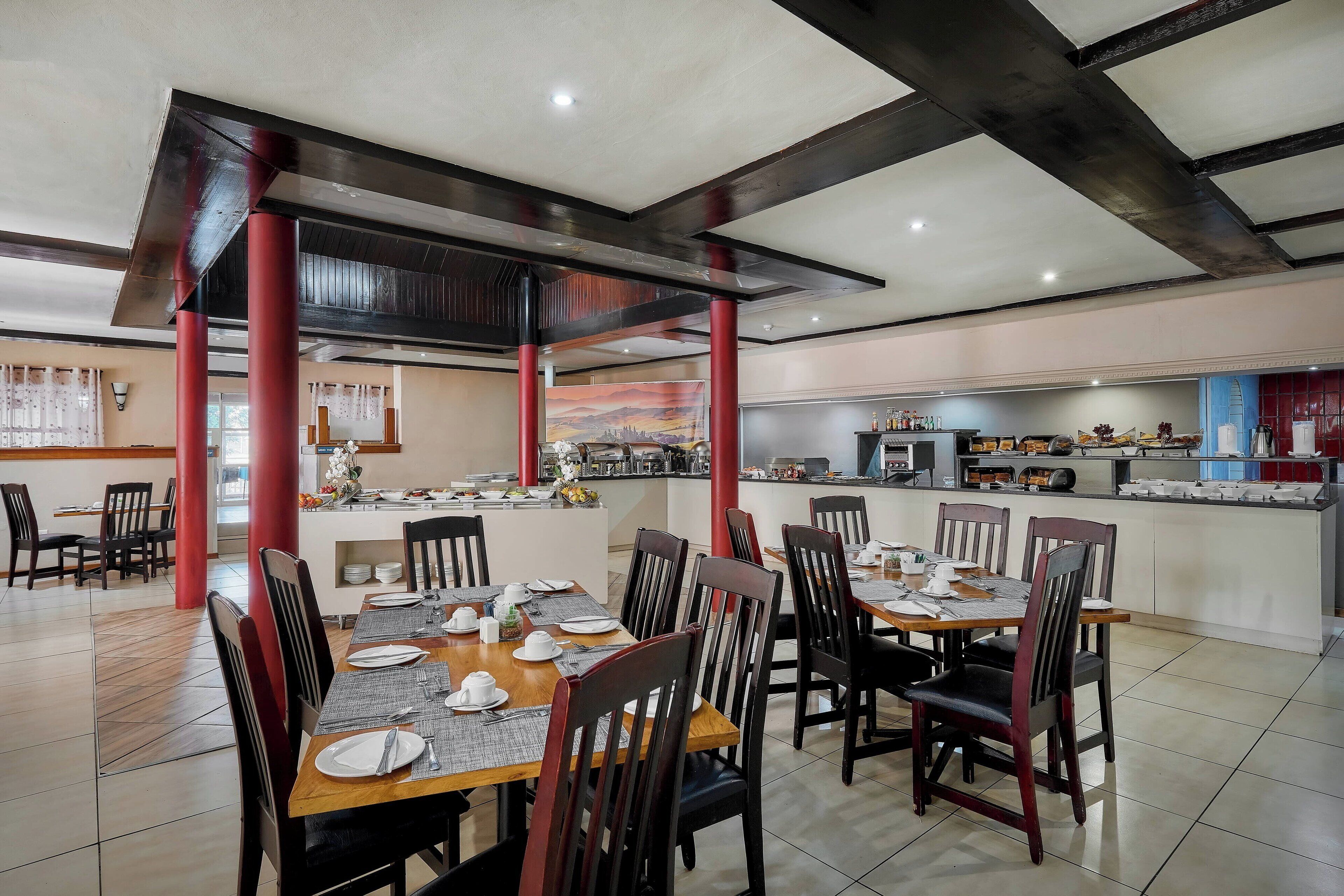 A spacious dining area with wooden tables and chairs, a central island with a kitchen in the background, and red pillars.