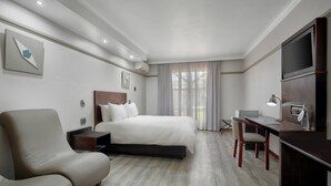 Quarto de hotel com cama, escrivaninha, cadeira, televisão e mesa de cabeceira.