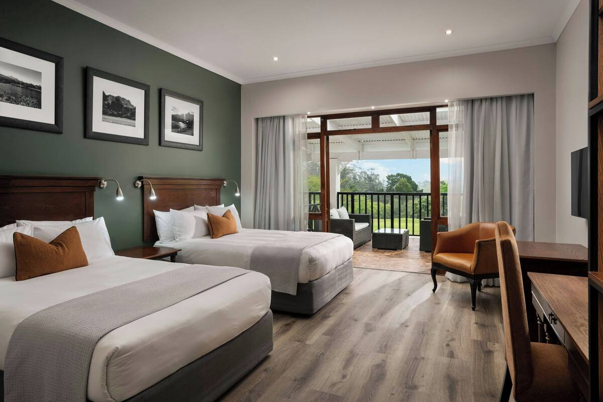 A hotel room with two beds, a desk, a chair, and a balcony with a view of the outdoors.
