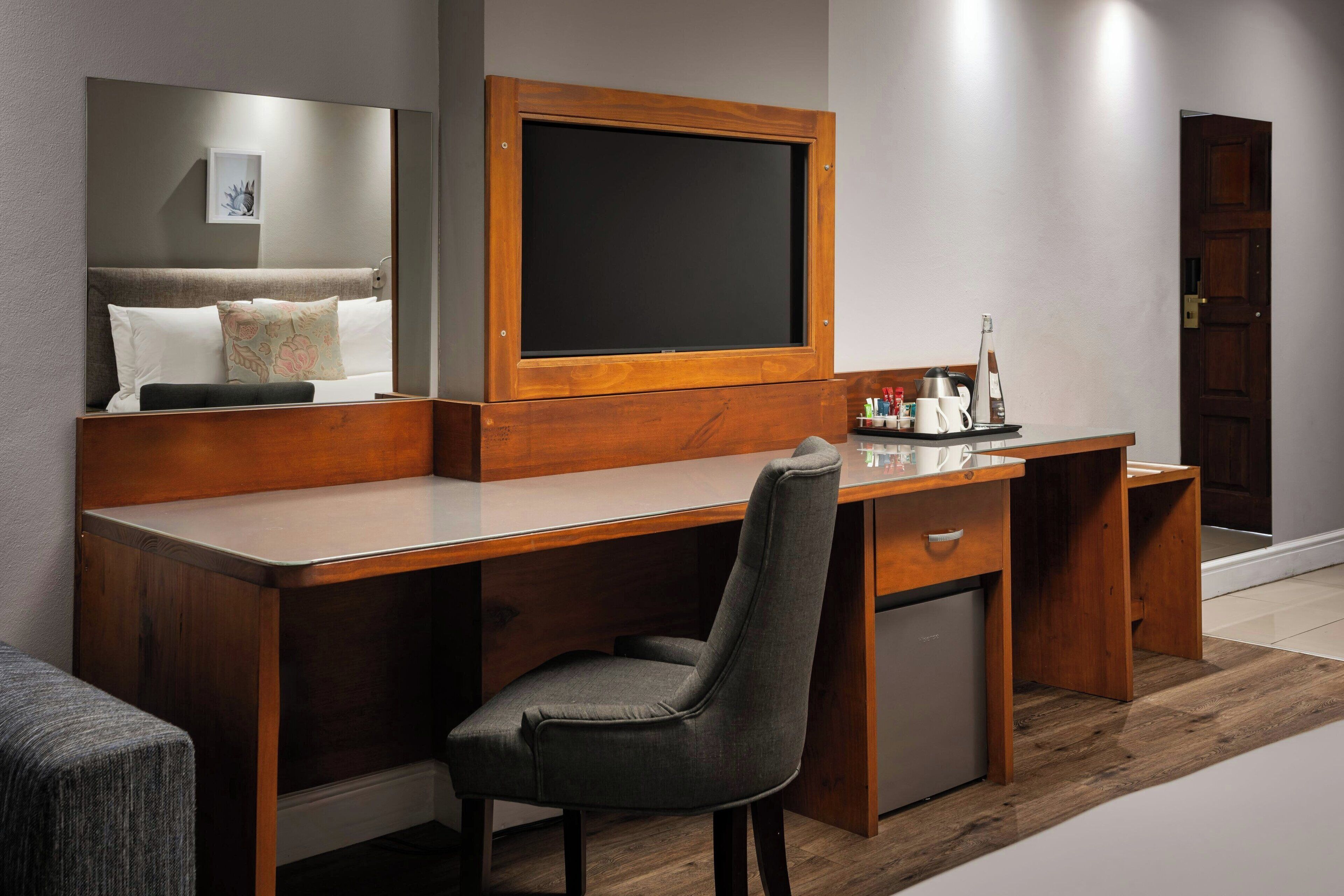Une chambre d’hôtel avec un bureau en bois, une chaise, un petit réfrigérateur, une table de chevet et un grand miroir.